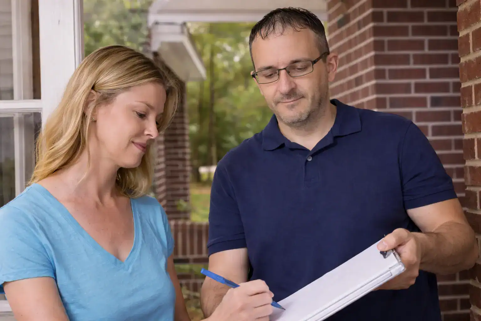 Service technician writing on clipboard while homeowner reviews paperwork outside brick house